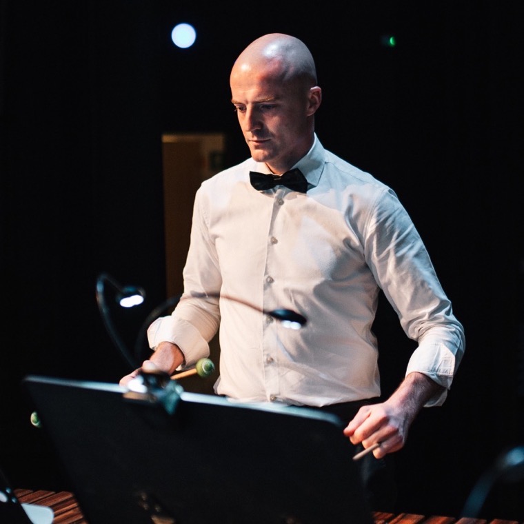 Dennis Vanhove speelt marimba in concertkledij met vlinderdas, op een donker podium.