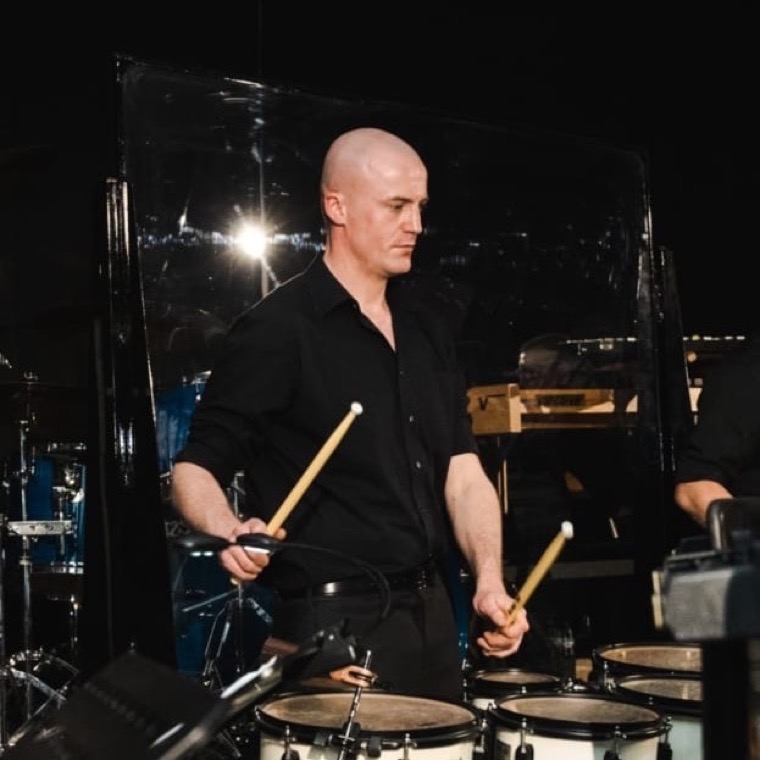 Dennis Vanhove speelt staand op een set trommels, in het zwart gekleed, met stokken in beide handen.
