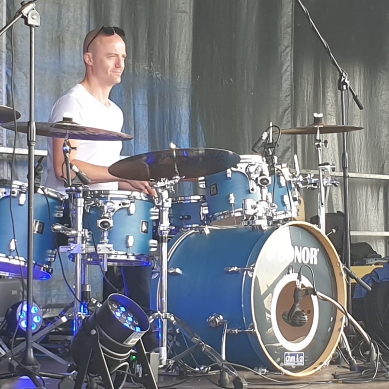 Dennis Vanhove achter een blauw drumstel op een openluchtpodium bij daglicht.