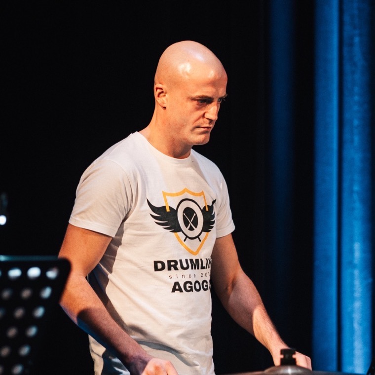 Dennis Vanhove met stokken in de hand, in een T-shirt met drumline-opdruk, voor een blauw podiumgordijn.