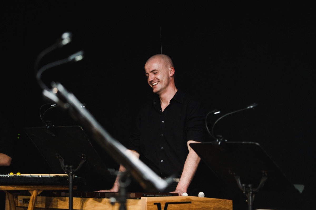 Dennis Vanhove lacht achter een marimba, in een zwart hemd, met lessenaars en microfoons rondom.